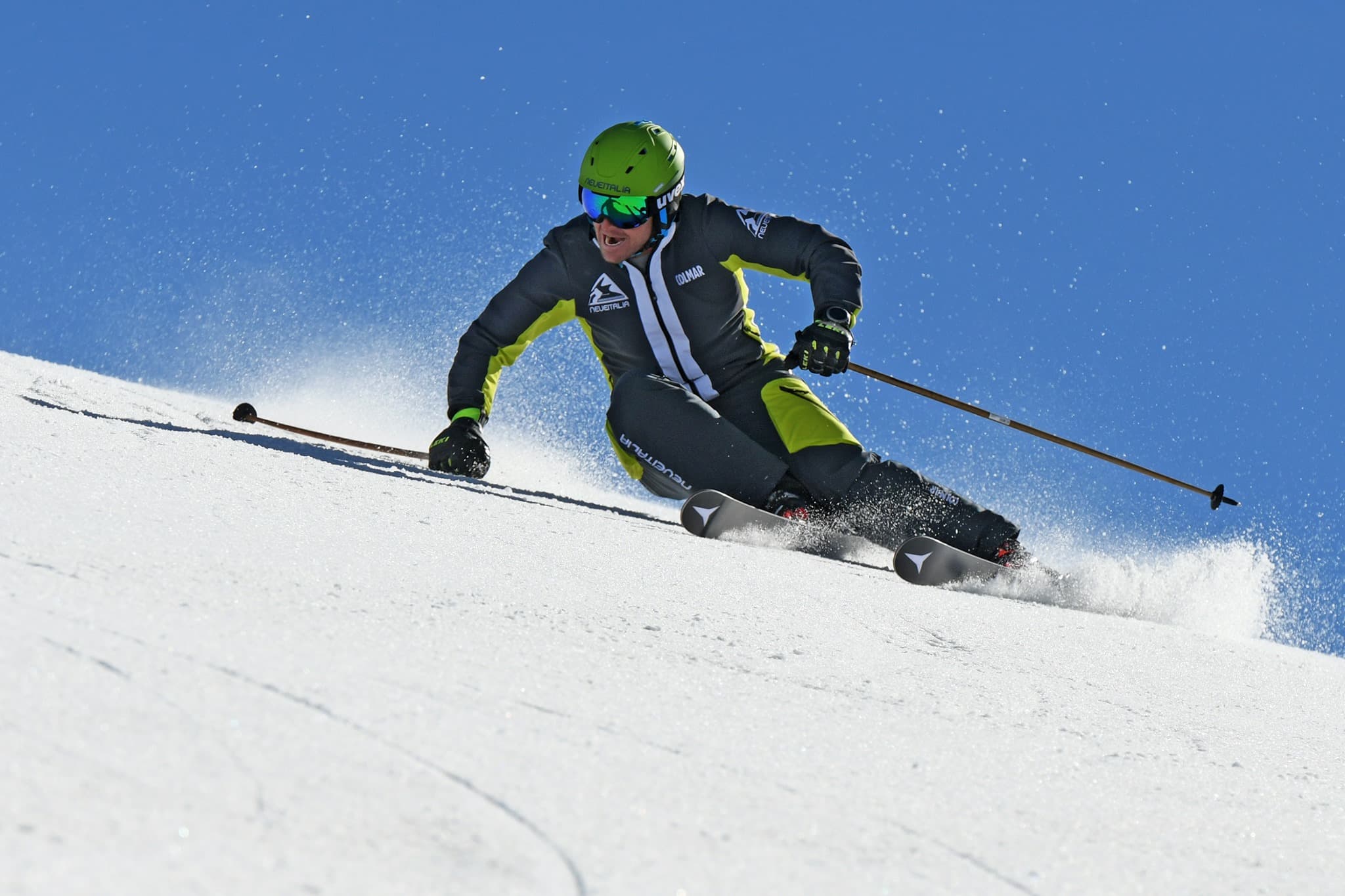 Sciatore con casco verde scende un pendio innevato sollevando neve sotto un cielo blu.