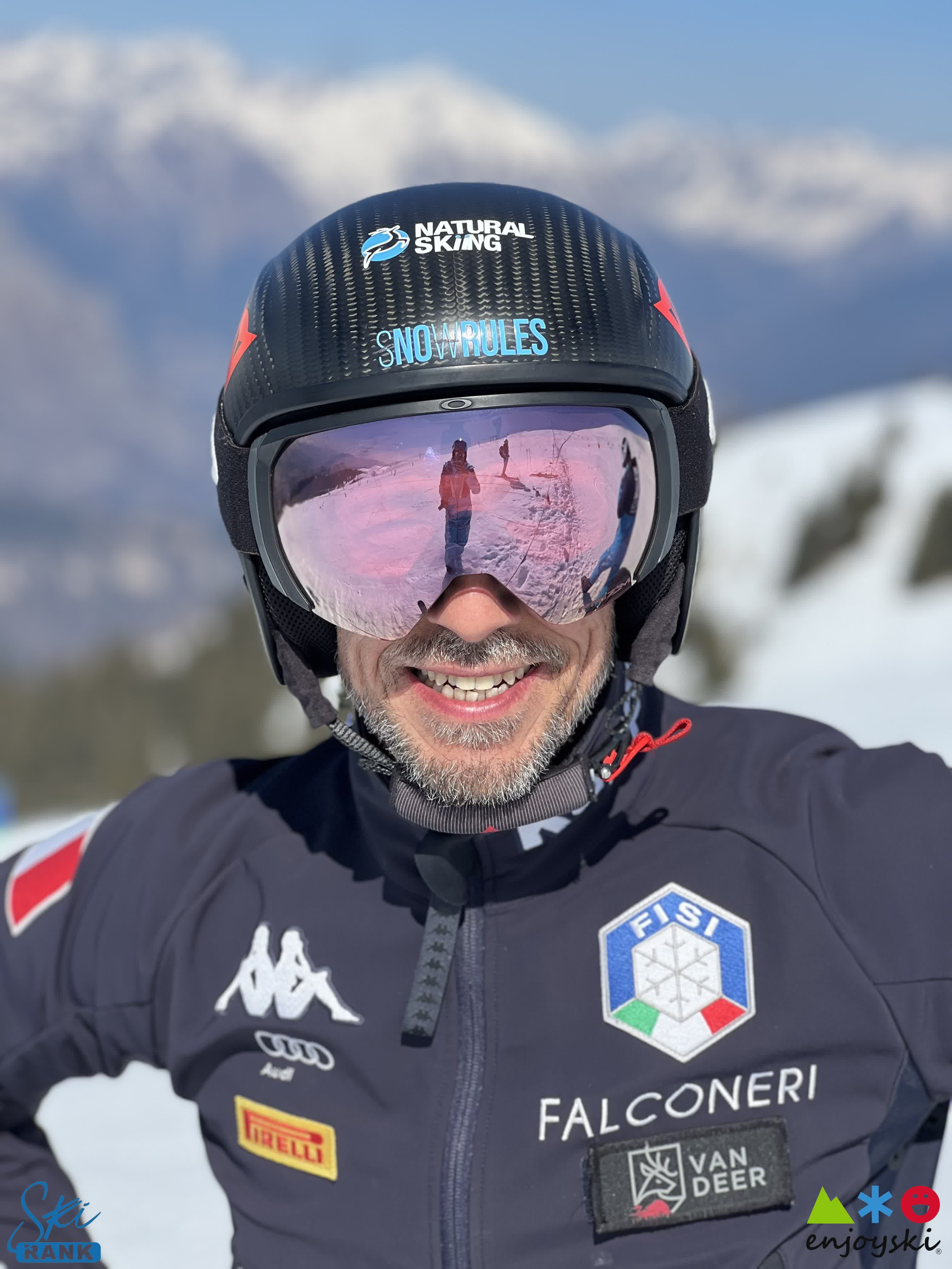 Sciatore sorridente con casco, maschera riflettente e giacca FISI davanti a montagne innevate.