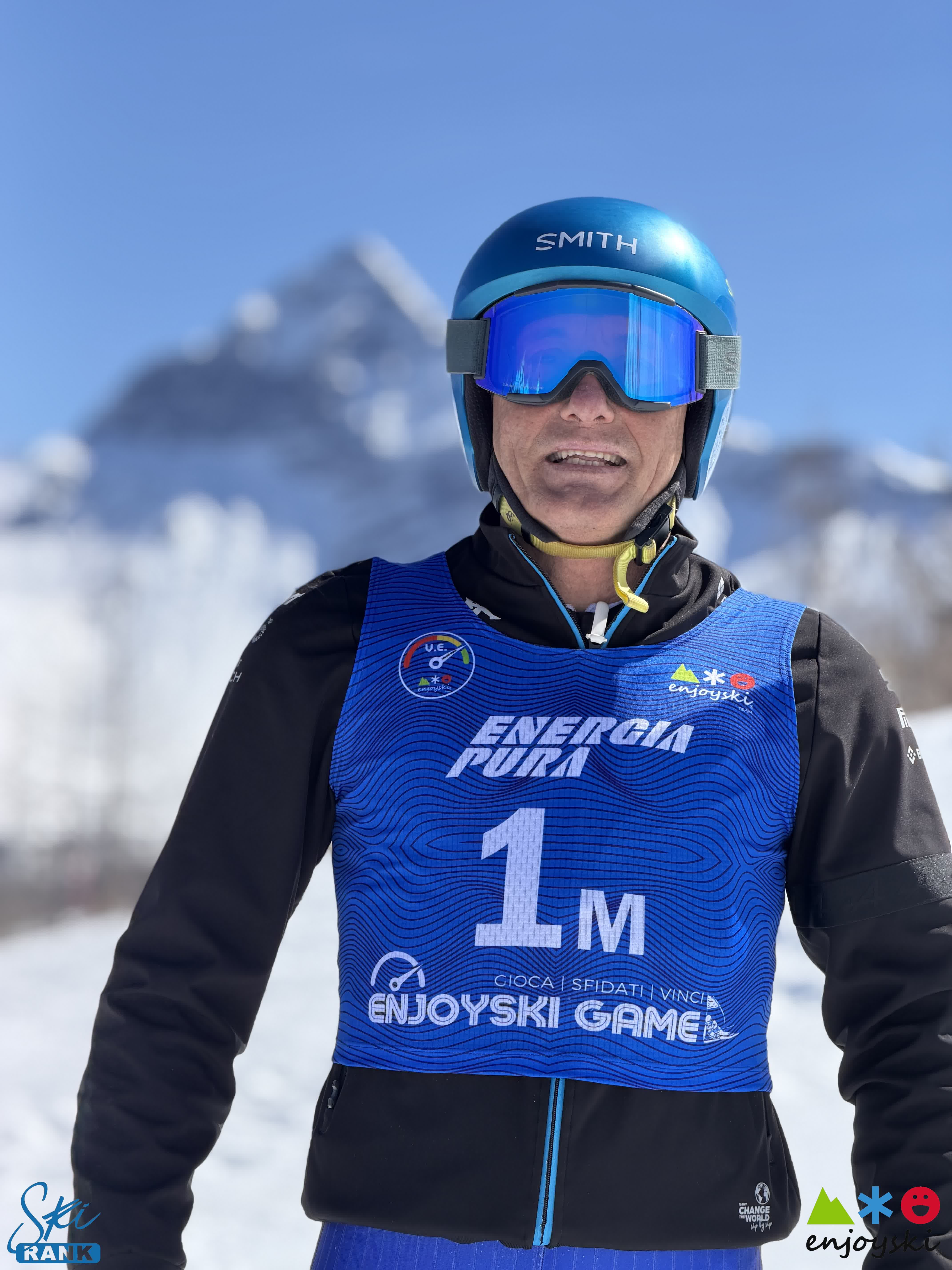 Sciatore con casco e pettorale blu numero 1M sorride davanti a maestose montagne innevate.