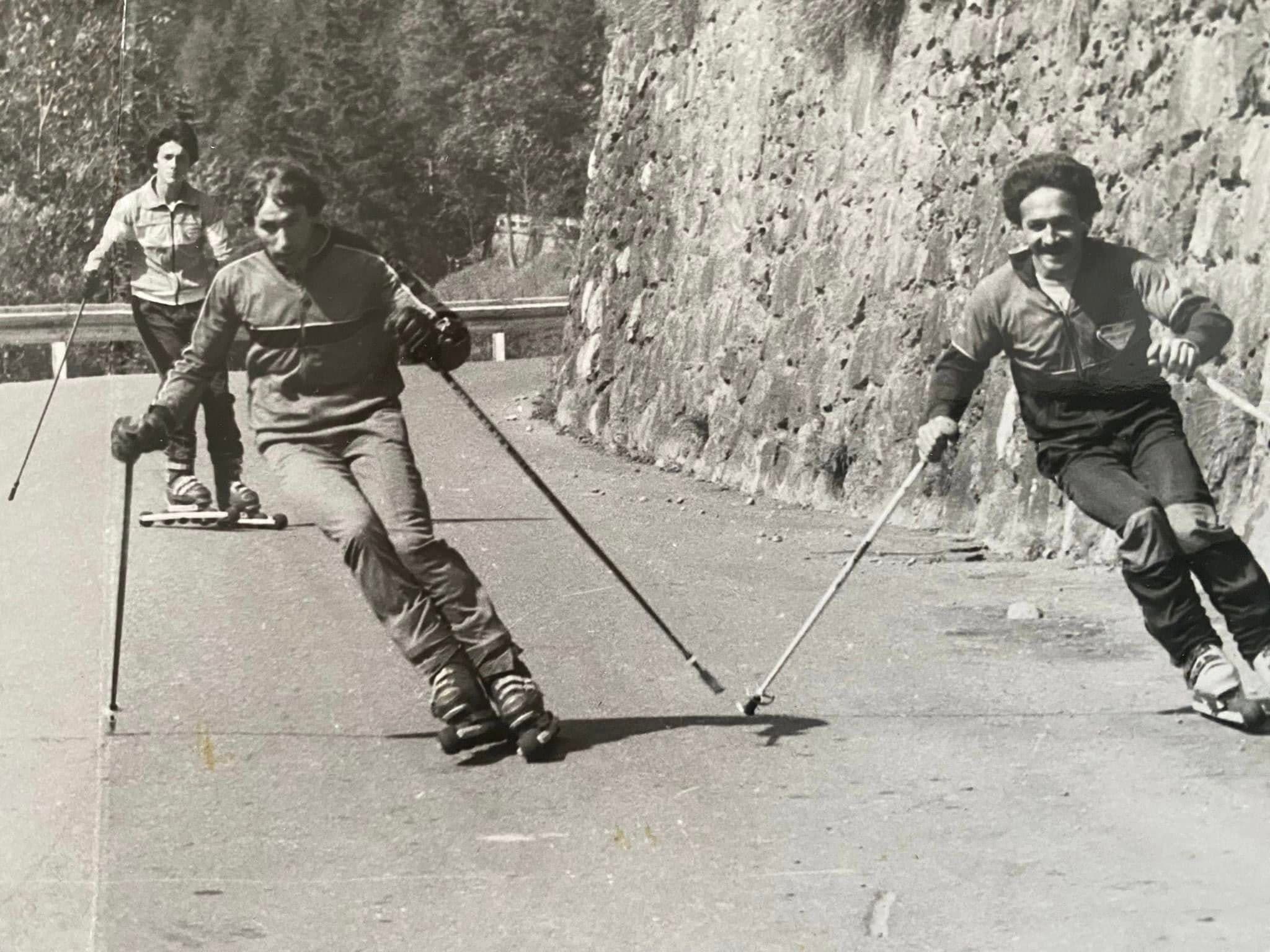 Tre uomini praticano sci a rotelle su una strada asfaltata di montagna con bastoncini.