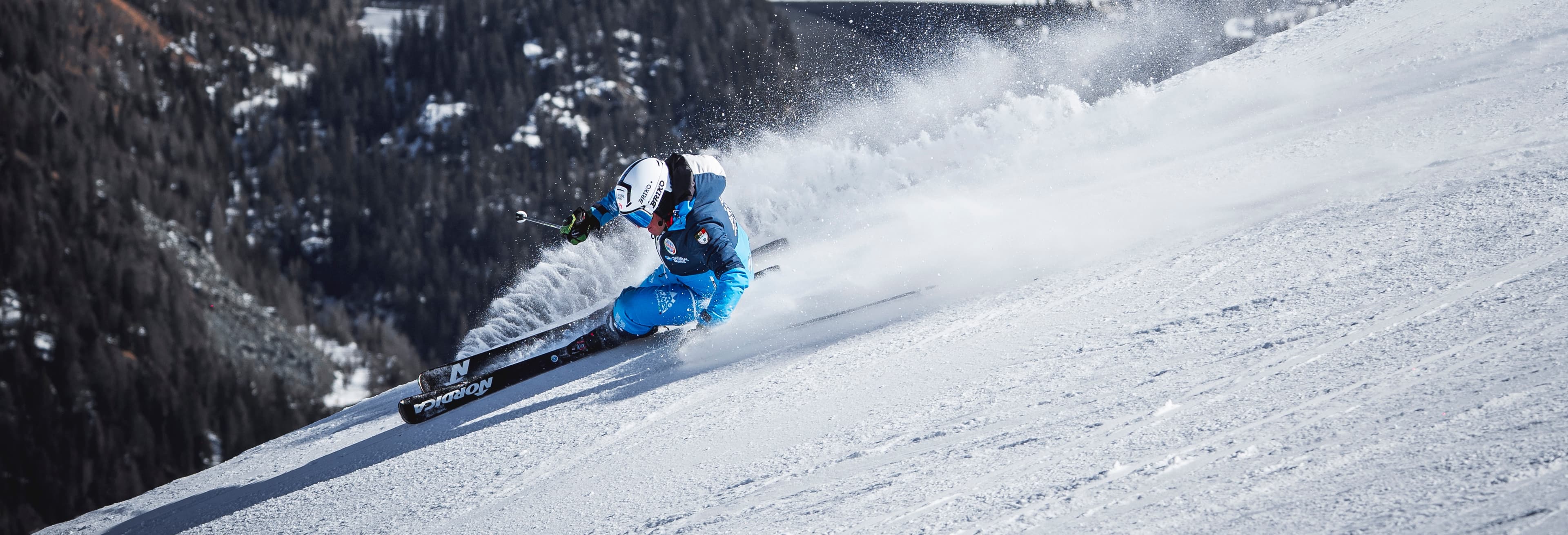 Sciatore in tuta blu che curva velocemente sulla neve sollevando una scia di spruzzi bianchi.