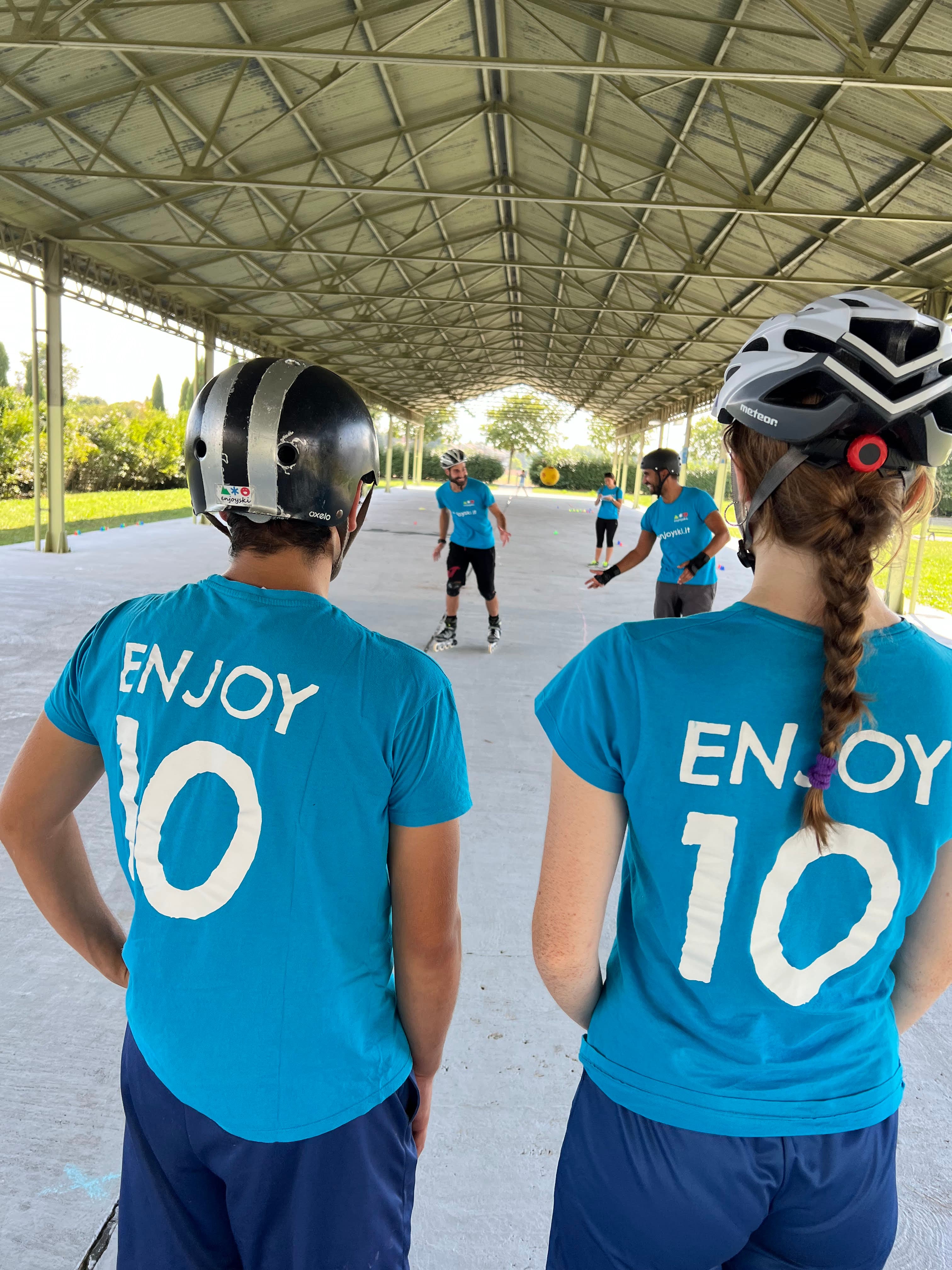Due persone con maglie 'ENJOY 10' osservano una partita di pattinaggio sotto una tettoia metallica.