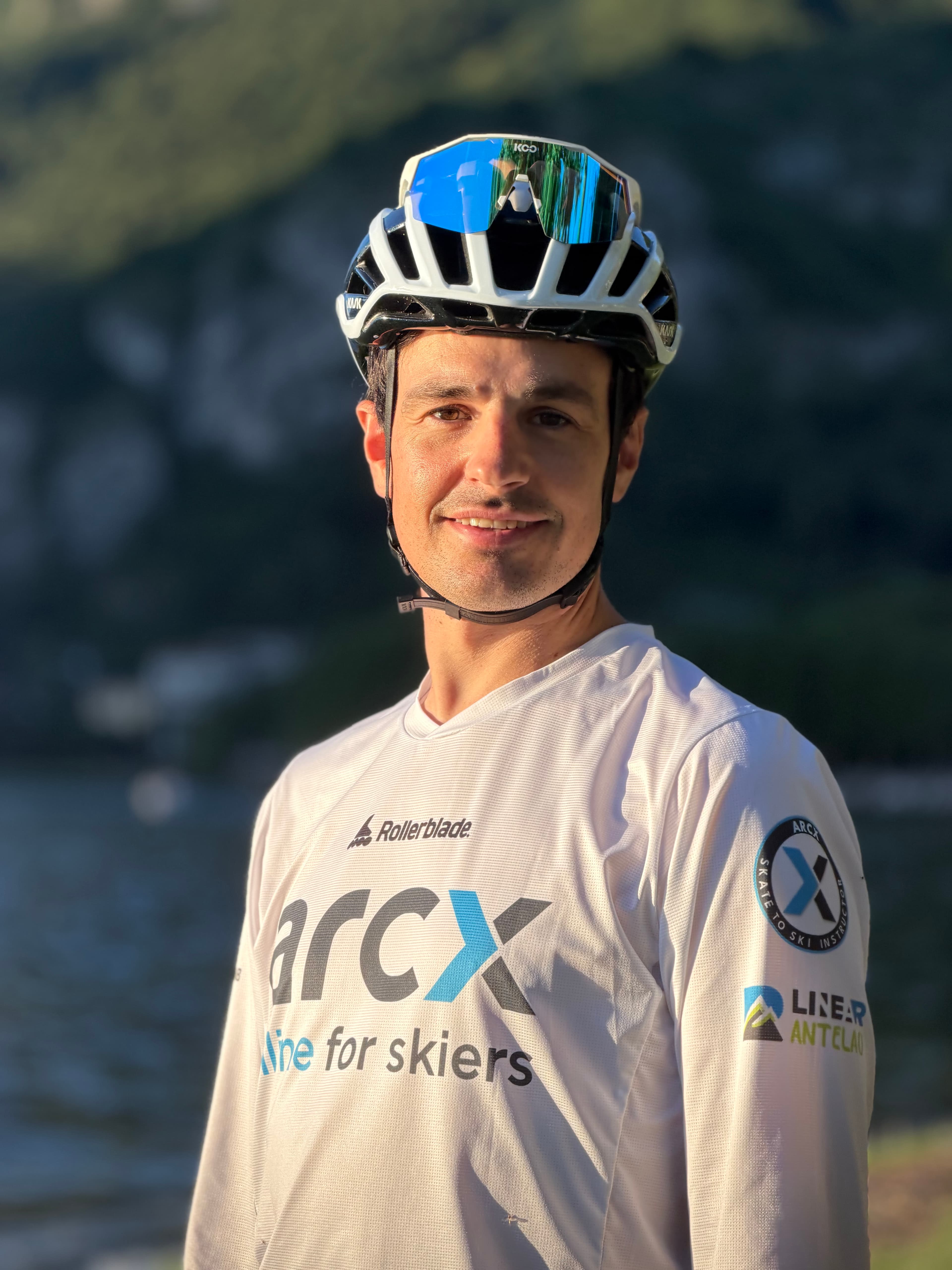 Uomo con casco da ciclismo e maglia bianca sportiva sorride davanti a un lago sfocato.
