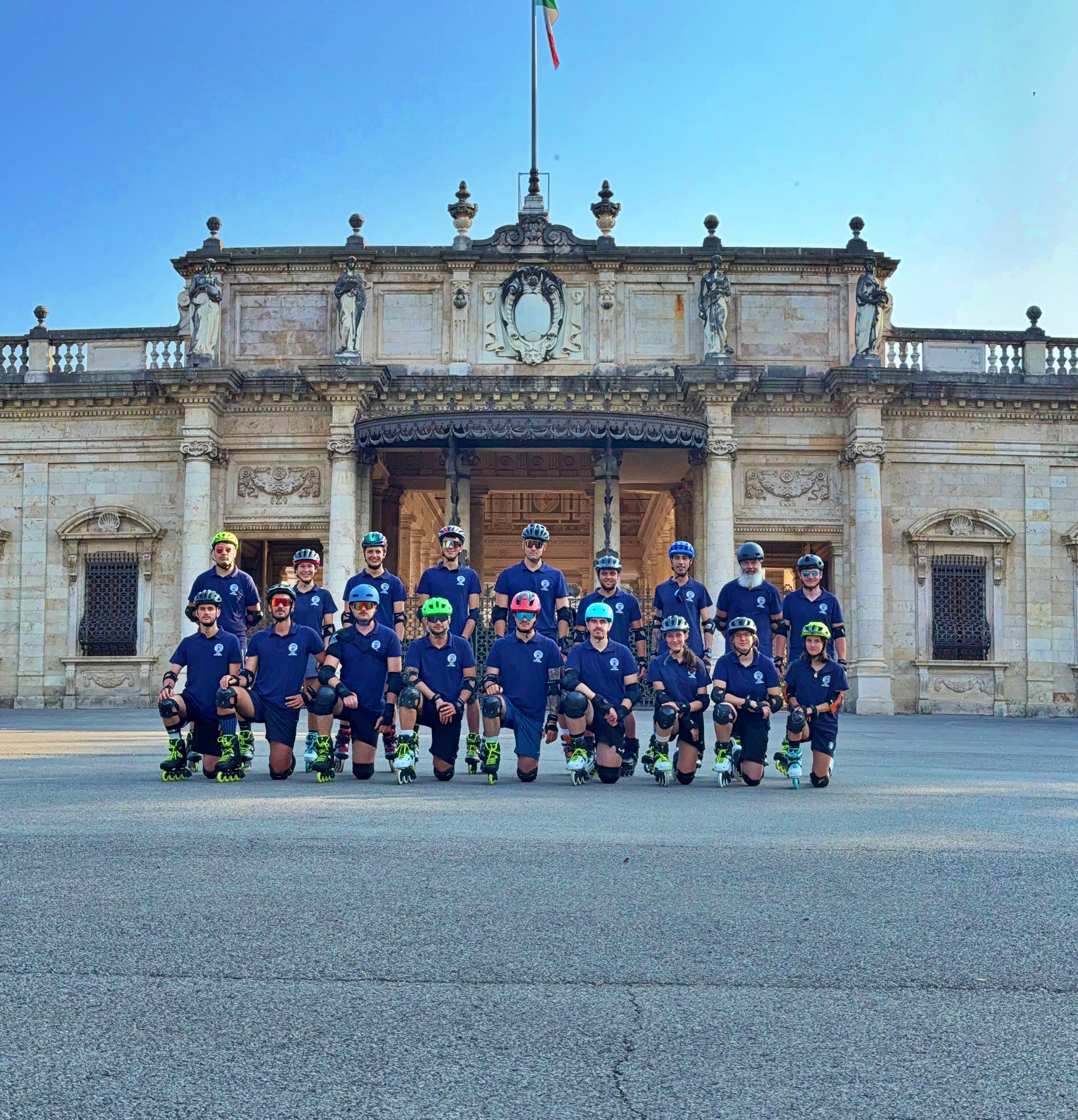 Gruppo di pattinatori in linea con caschi colorati posa davanti a un imponente palazzo storico.