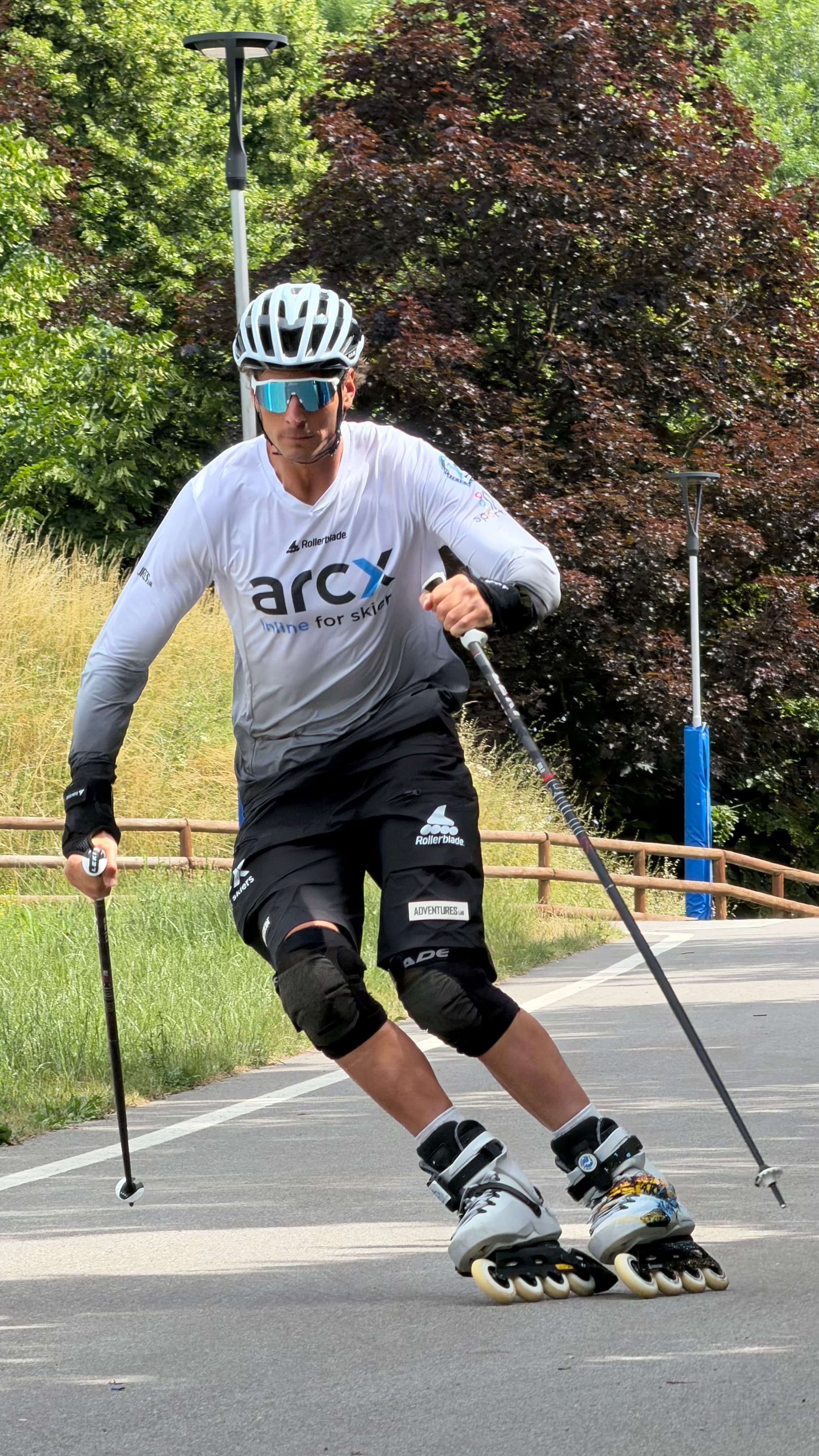 Pattinatore in linea con casco e bastoncini da sci si allena su un sentiero asfaltato.