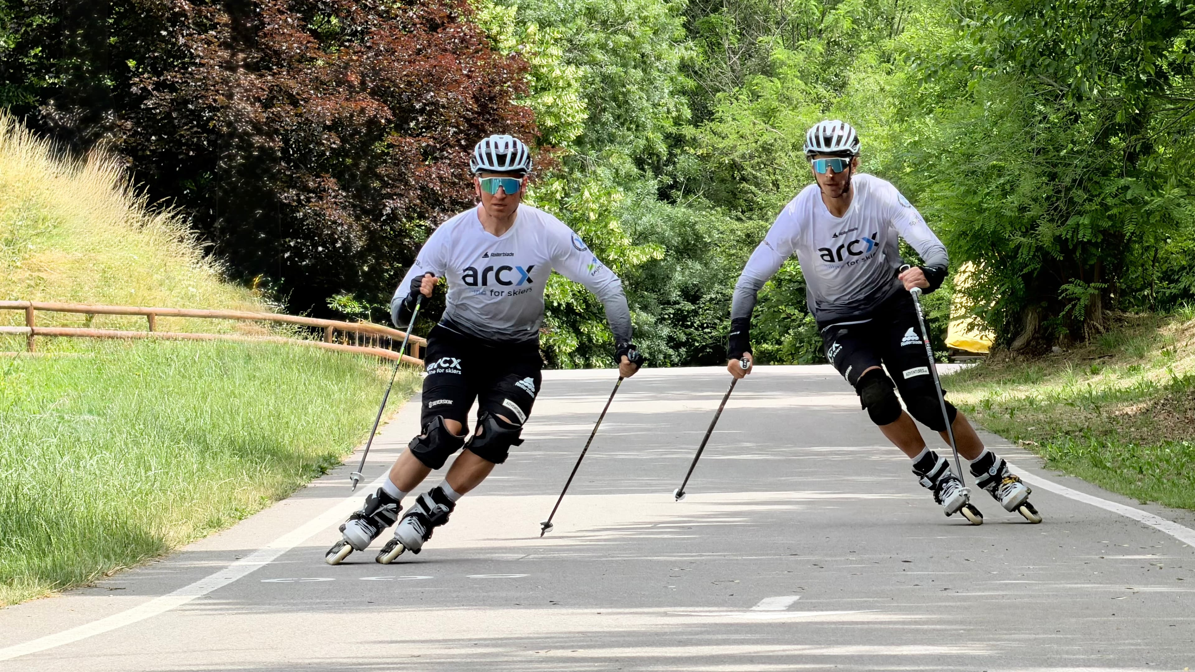 Due uomini con caschi e bastoncini fanno rollerblade su una strada asfaltata circondata da alberi.