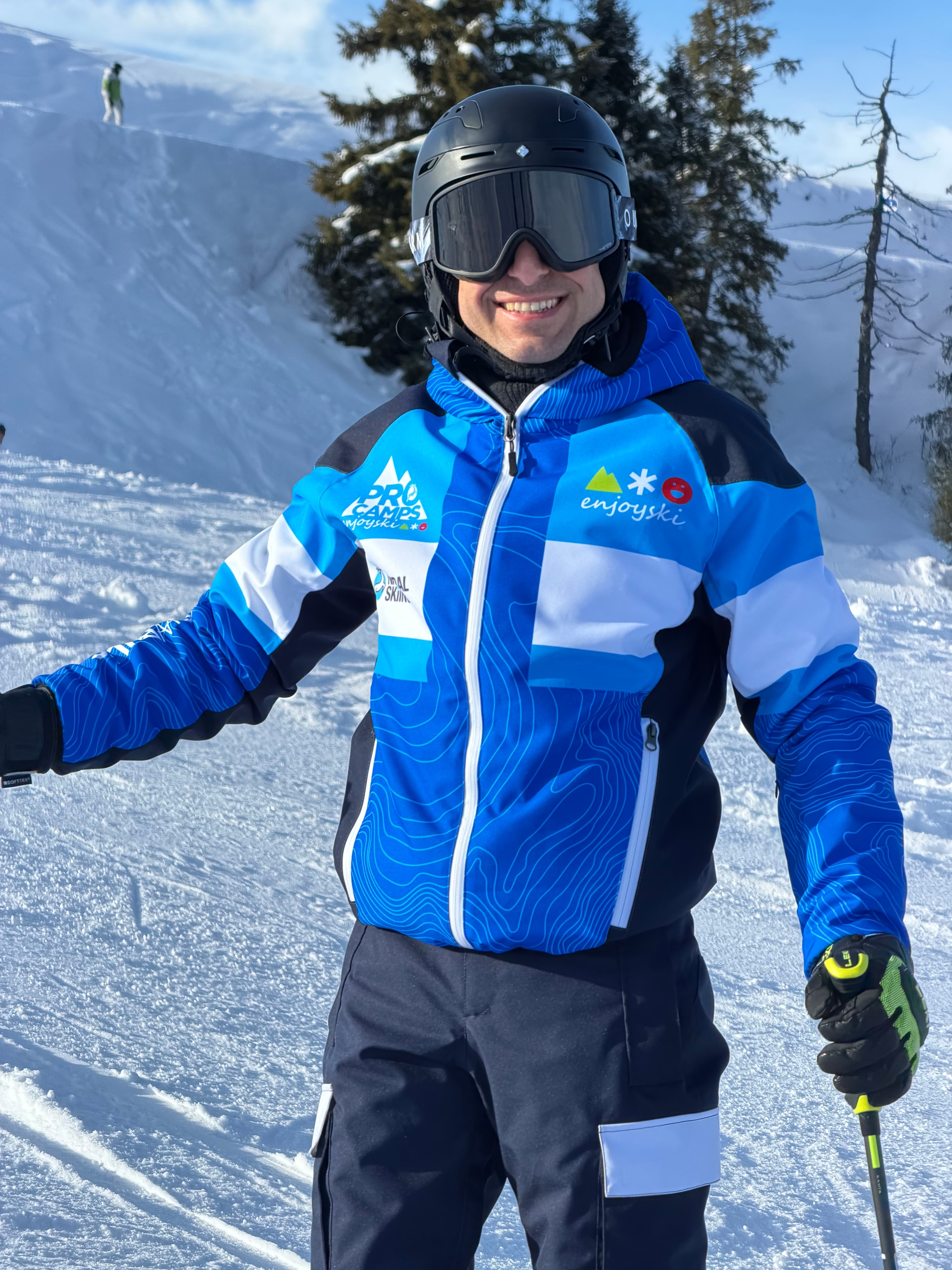 Sciatore sorridente con casco e giacca blu e bianca su una pista da sci innevata.