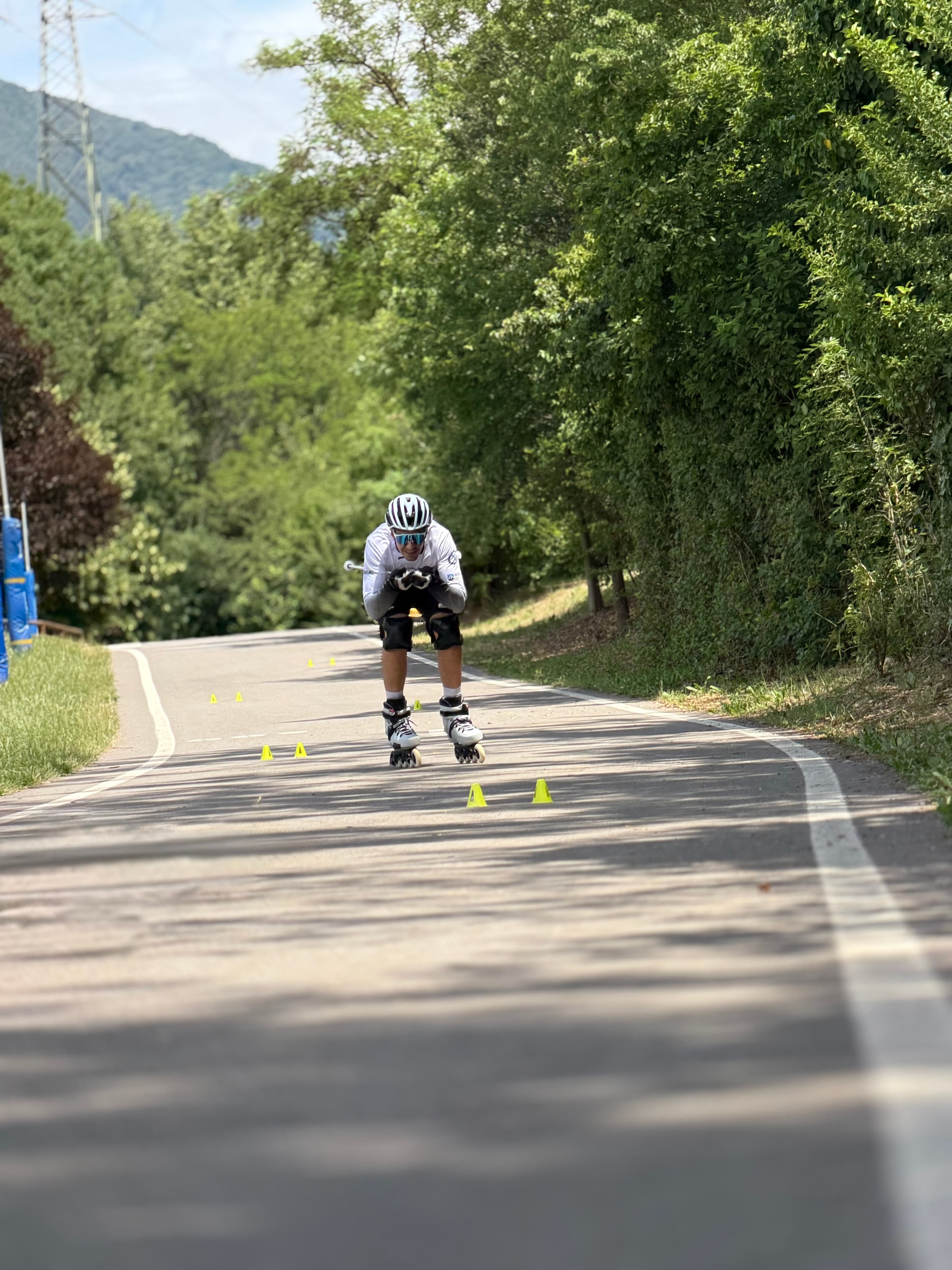 Pattinatore in linea in posizione aerodinamica scende tra birilli gialli su una pista ciclabile.